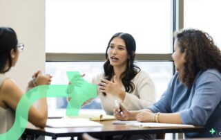 Three professional women engaged in a collaborative discussion at a table in a bright office setting, with notebooks and pens in front of them. One woman speaks while the others listen attentively. A translucent green arrow graphic and the Discflow logo are overlaid on the image.
